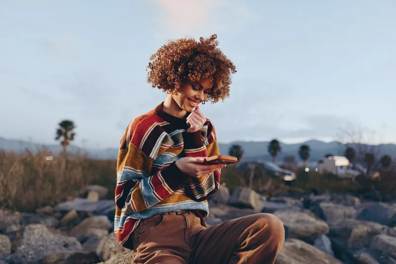 Woman lifestyle, outdoors in nature, curly hair woman sitting on rocks, looking at phone screen, warm sunset light, distant mountains and calm landscape, relaxed confident mood