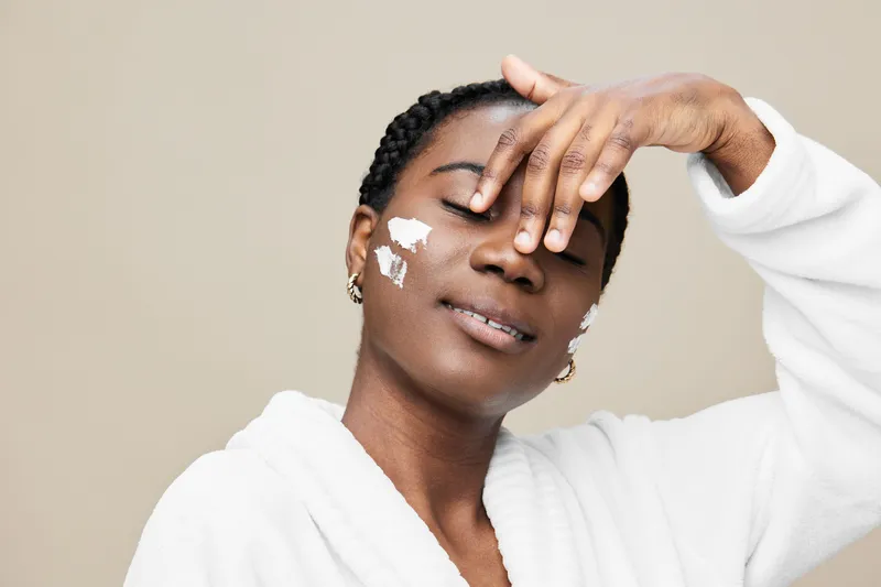 Young Black woman applying facial cream, showcasing healthy skincare routine with a peaceful expression in a neutral toned background