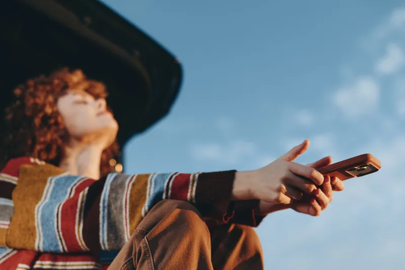 Woman lifestyle outdoors, hands holding smartphone in the open air, wearing a hat and looking toward blue sky. Casual communication moment, sunny day portrait for travel planning.