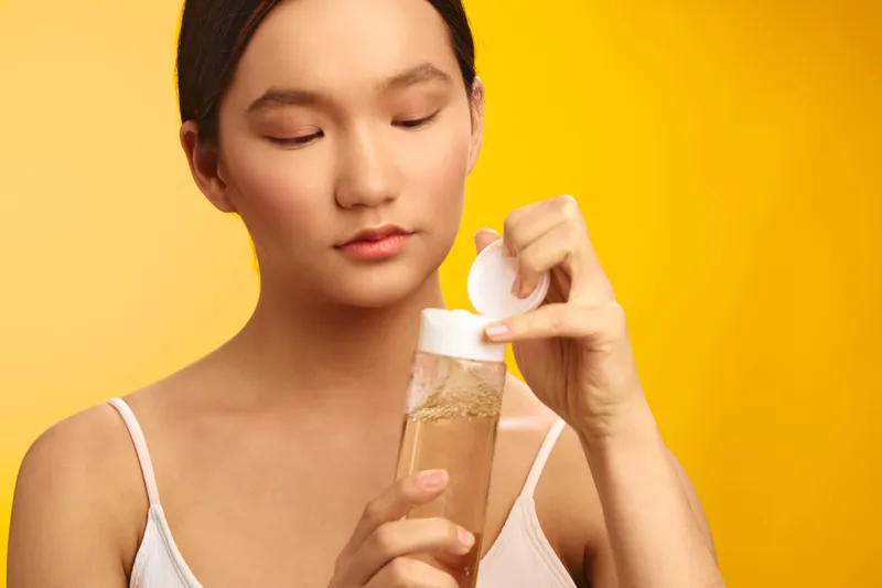asian woman pours liquid into a bottle in a bright studio. She concentrates on her task, conveying a calm, mindful skincare moment with a clean, simple setup.