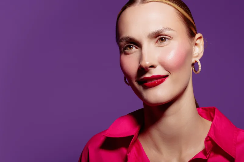Smiling woman with red lipstick and pink shirt poses against purple background. Close-up portrait with hoop earrings and natural makeup. Concept of beauty, fashion, and confidence.