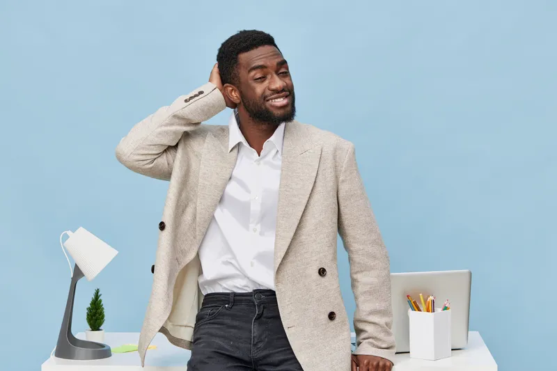 Confident smiling man with dark skin, wearing beige blazer and white shirt, standing in modern office space with light blue background, in casual pose, showing relaxed attitude, office ambient, busine