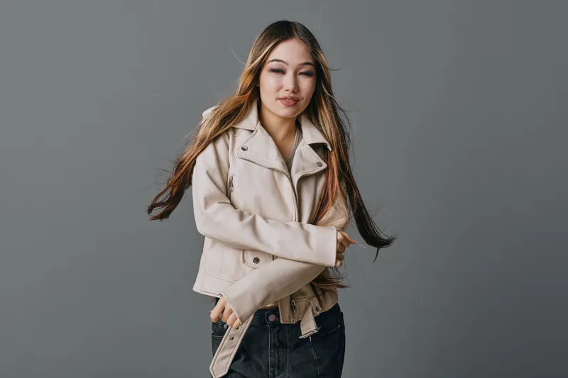 Woman with long hair wearing beige jacket and black jeans standing on gray background looking at camera with natural makeup and relaxed pose for portrait or fashion concept.