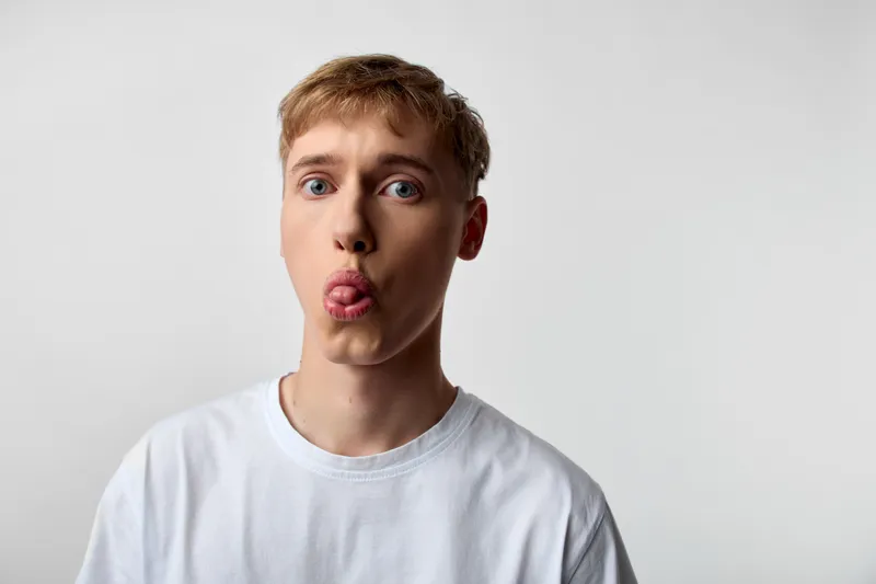 Young man with tongue out, playful expression wearing a white tee against a simple light background, casual fun, suitable for humor, lifestyle, and social media branding projects