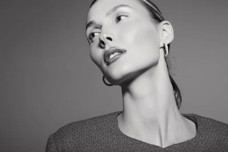 Portrait of a young white woman with pale skin and slicked back hair wearing a dark top. Studio lighting highlights her sharp features and elegant earrings in black and white style.