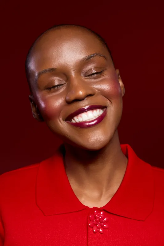 Close-up of happy African woman with dark skin and shaved head smiling with closed eyes wearing red top against red background. Concept of beauty, confidence, and natural skincare.