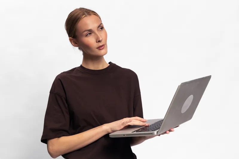 Young woman in casual dark shirt holding laptop and looking away thoughtfully isolated on white background. Concept for technology, remote work, analysis, and concentration.