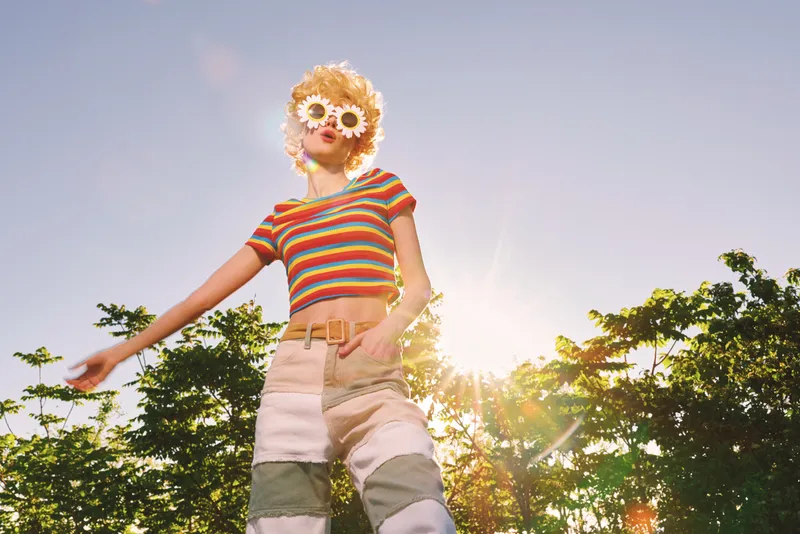 A stylish woman in a striped crop top and wide shorts stands outdoors in bright sunlight, wearing playful sunglasses and posing confidently. Retro film color adds warmth to the vibrant outdoor scene,