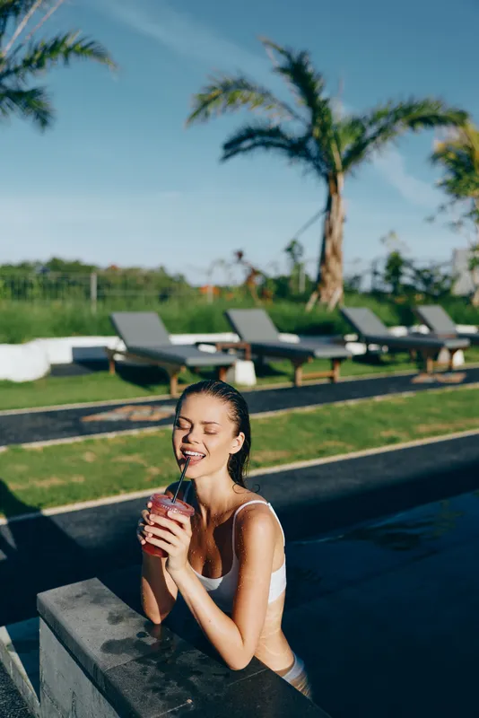 A woman in a white swimsuit enjoys a refreshing drink by a poolside in bright sun, capturing a relaxed lifestyle moment. The resort setting features palm trees, lounge chairs, and clear blue skies.