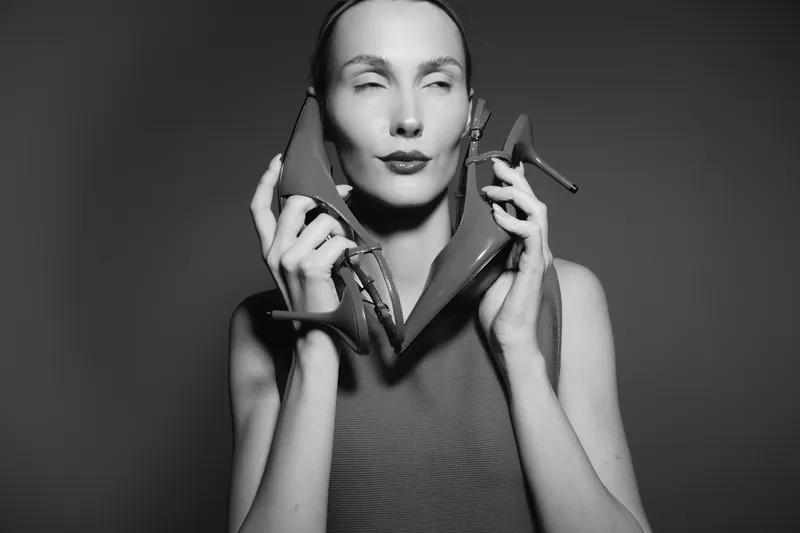 White woman with fair skin holds high heel shoes near face with closed eyes, wearing sleeveless top, studio portrait in black and white.