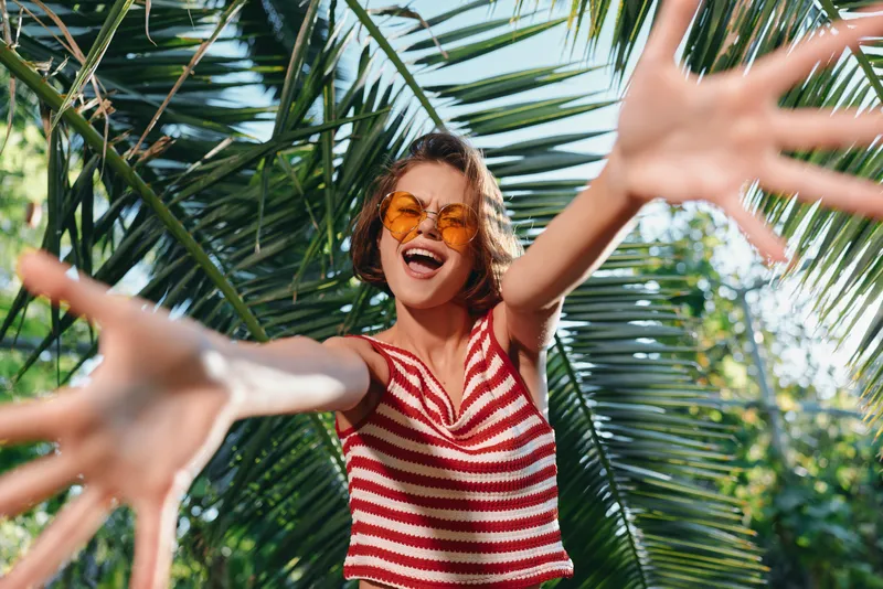 A joyful person wearing a red and white striped top reaches toward the camera in a sunny outdoor setting. Palm leaves frame the scene, creating a carefree, energetic mood.