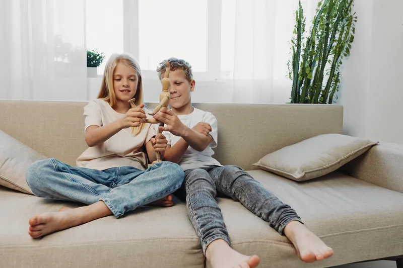 Two children sit barefoot on a beige sofa playing with dolls in a bright living room, natural light creating a relaxed and cozy atmosphere for family time.