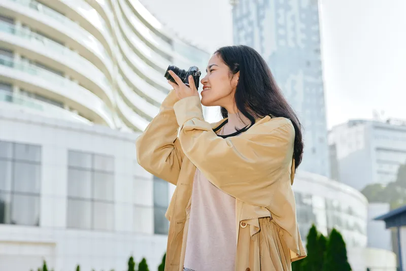 Woman photographer captures cityscape with a compact camera, standing outdoors in an urban environment, wearing a beige jacket and casual attire, focused on street photography, embracing urban life