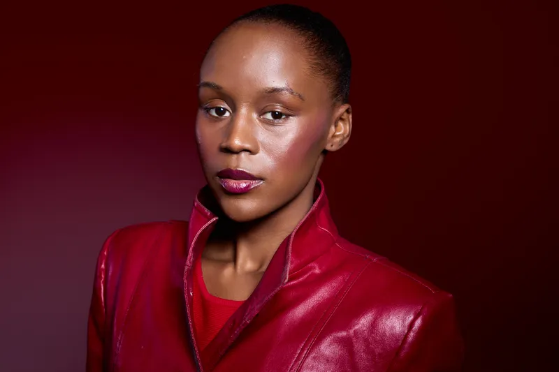 Confident African woman with dark skin wears a red leather jacket and red top, posing against a deep red background. Concept of fashion, beauty, and elegance with strong color contrast.