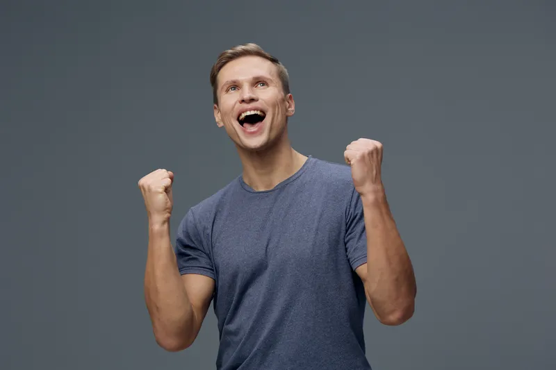 Joyful man celebrating success with fists raised in excitement, casual blue T-shirt, joyful expression, positive mood, plain gray background, studio shot, human emotions.