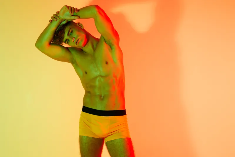 Fit man stretching arms standing, shirtless muscular fitness model posing against color background in sports brief underwear for exercise health stretching concept outdoors.