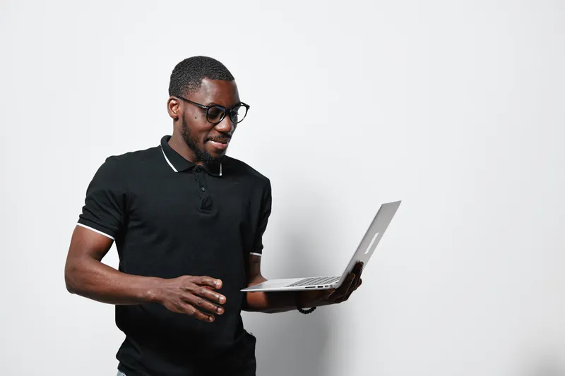 African American man with dark skin and glasses using laptop for work or study in casual black polo shirt on white background. Technology, communication and digital device.
