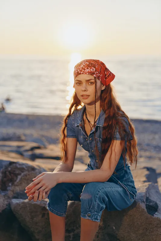 Lifestyle woman with modern boho-western denim street style sitting on rocks by the ocean during golden hour in warm film color tones, relaxed and contemplative mood.