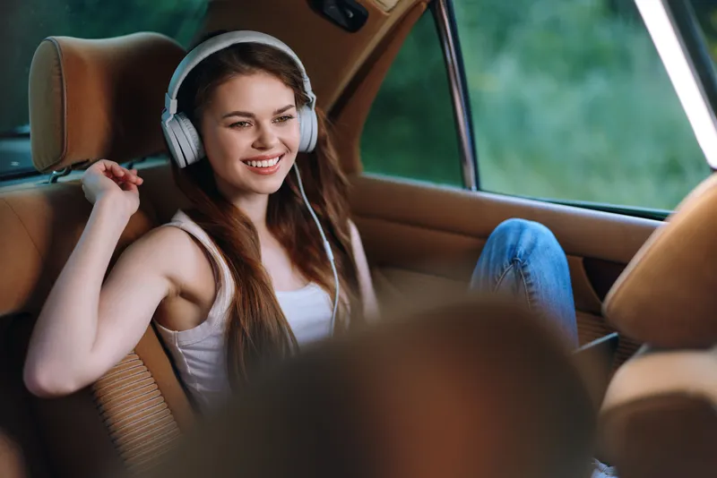 Young woman enjoying music inside a car, wearing headphones, smiling happily. The soft green background enhances a relaxed and cheerful mood.
