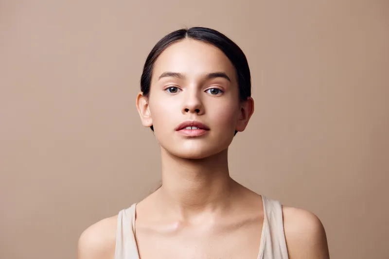 Natural beauty portrait of a young woman with a calm expression, wearing a beige top, against a soft neutral background, highlighting fresh skincare and simplicity