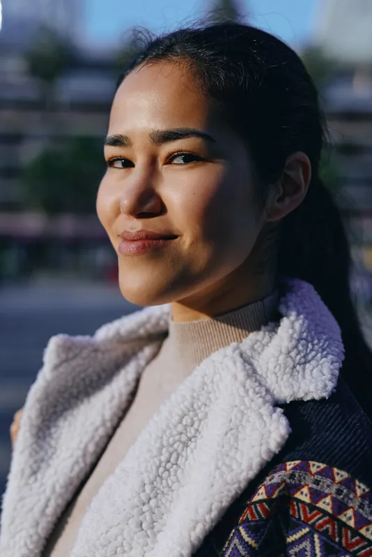 Woman smiling outdoors wearing a warm fleece jacket and scarf in sunlit urban light, casual fashion mood, natural candid moment with soft textures and cozy appeal