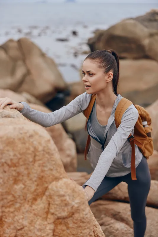 Young woman climbing rocks by the sea, demonstrating determination and focus in activewear and a backpack, showcasing an adventurous lifestyle in a serene environment