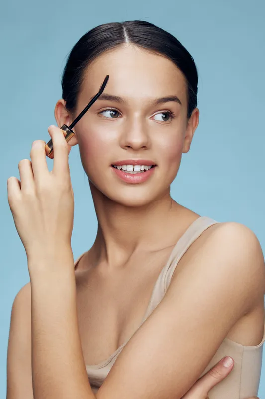Young woman applying eyebrow gel, showcasing natural beauty with a soft smile against a pastel blue background, ideal for beauty and skincare concepts