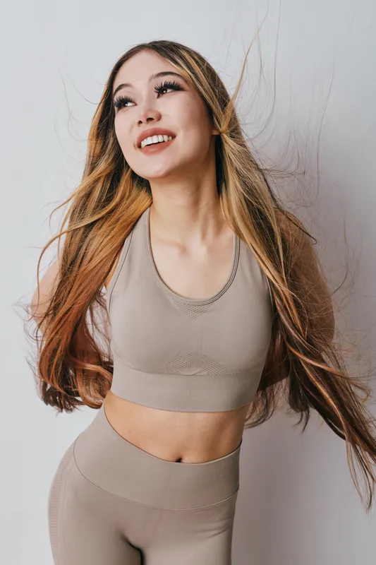 Young woman with long hair wearing beige sportswear smiling and posing on white background. Fitness, beauty and healthy lifestyle concept with natural look and positive expression.