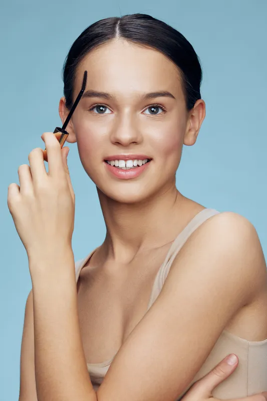 Young woman applying eyebrow gel with a smiling expression against a soft blue background, emphasizing beauty and self care concepts in a fresh, modern style