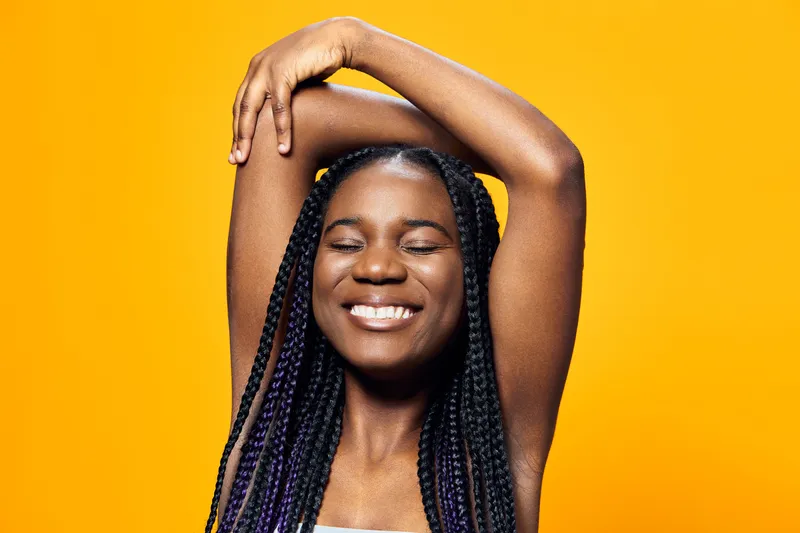 Happy young woman with braided hair expressing joy against a vibrant yellow background, showcasing positivity and self confidence in a casual style