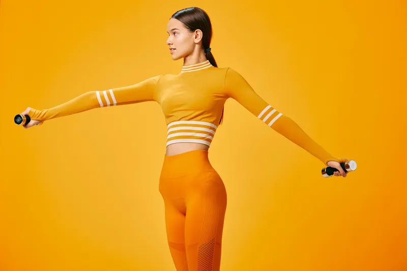 Young woman fitness model in matching orange workout set performs lateral arm raises with small dumbbells against a seamless vibrant orange background, athletic posture and focused expression.