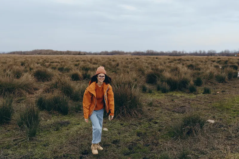 Woman lifestyle outdoors walking in grassy field, orange jacket and knit beanie, calm smile, cloudy sky, casual hiking, countryside nature, long shot full body, soft winter light, boots