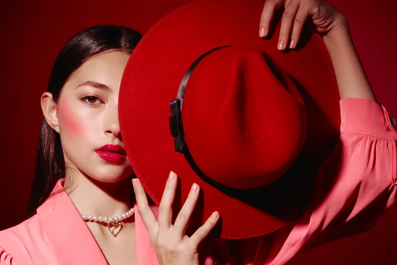 Beautiful woman with perfect makeup and a vibrant red hat holds the hat partially covering her face against red background. Fashion concept with creative beauty styling and elegant pose.