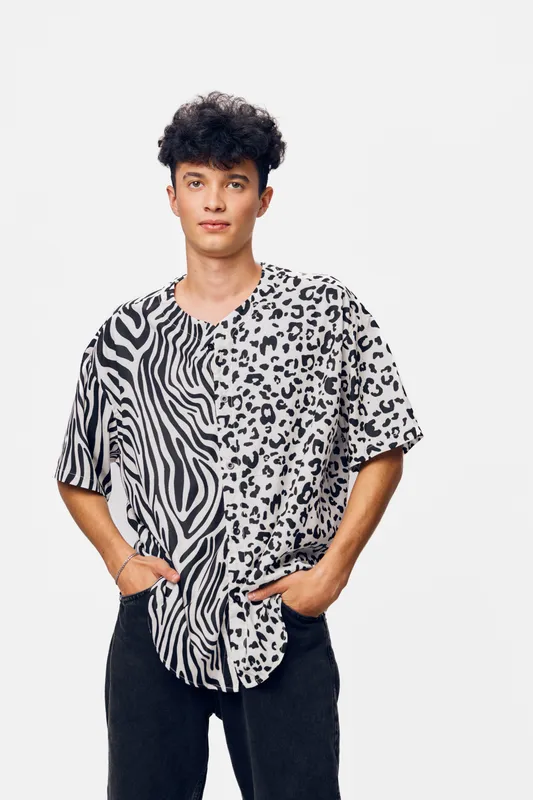 Young man wearing a black and white animal print shirt stands confidently with hands in pockets against a plain white background, showcasing modern casual style.