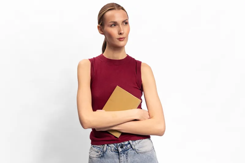 Young woman in sleeveless red top holding a notebook, looking away thoughtfully. Casual style with light blue jeans and simple hairstyle on white background. Concept for planning and focus.