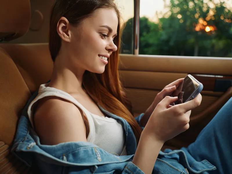 Young woman smiling while using smartphone in a cozy car, wearing casual denim jacket and white tank top, capturing joyful moments in a summer setting.