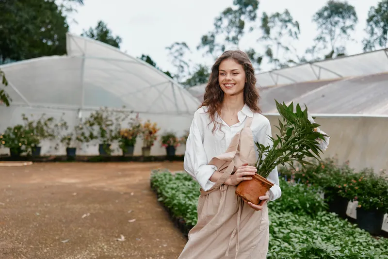 Young woman smiling in a greenhouse, wearing a white shirt and beige overalls, holding a potted plant, conveying joy and connection with nature