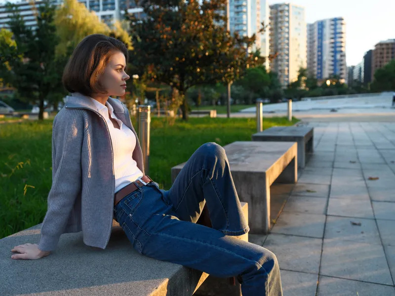 A confident woman sits on a concrete bench in an urban park during golden hour. She wears a light jacket and jeans, exuding casual style and calm confidence.