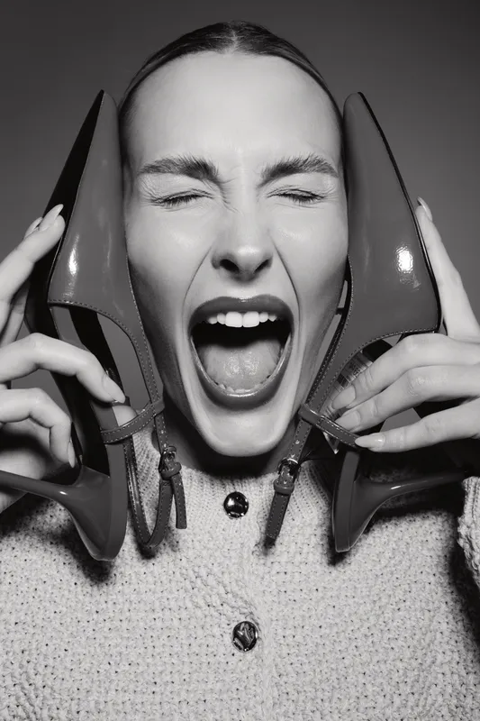 Young Caucasian woman with fair skin holding high heel shoes near face, eyes closed and mouth open. Concept of fashion, emotion, and beauty in black and white portrait.