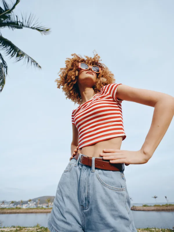 Young woman stands outdoors in a red and white striped crop top and high waisted jeans, posing confidently under clear sky with palm trees and seaside backdrop.