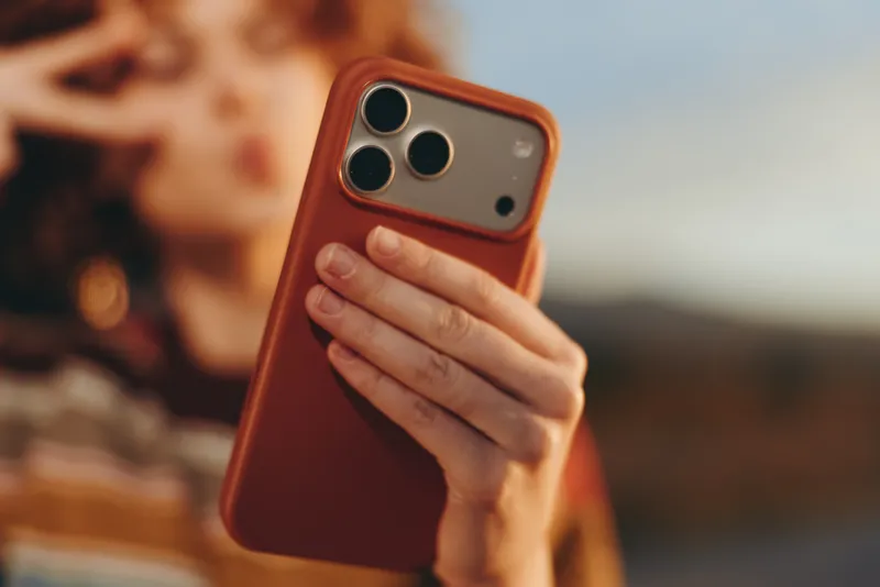 Woman lifestyle holding smartphone close to face outdoors, warm sunset light, soft focus background, hand covering camera module, casual travel portrait, modern communication and everyday tech
