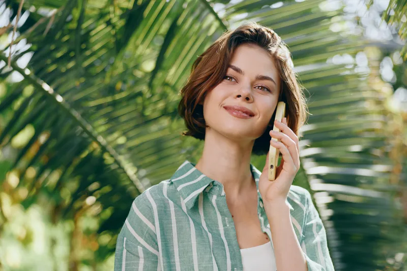 Woman smiles while talking on a smartphone outdoors, framed by lush palm leaves and bright sunlight, wearing a striped green shirt.