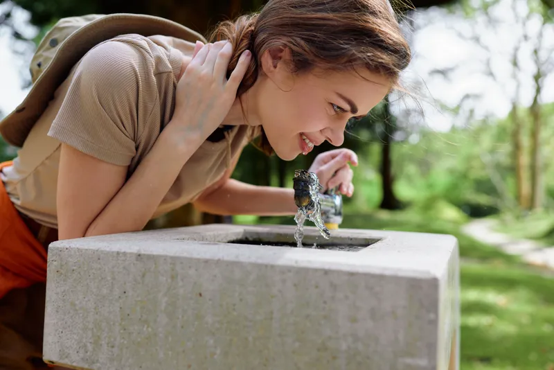Woman drinks water from outdoor stone fountain in park, leaning over with backpack and smiling, hydrating on a sunny summer day in nature, closeup portrait with green background