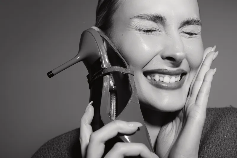 Smiling Caucasian woman with fair skin holds high heel shoe near face, eyes closed and happy. Concept of fashion, beauty and joyful emotion in black and white.