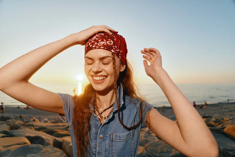 smiling woman with bandana enjoying sunset on rocky beach, casual outfit, natural light, serene expression, warm colors, summer vibe