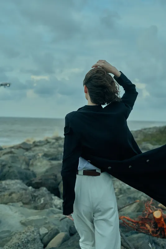 Individual in a dark coat stands with back to camera on a rocky coast, wind catching flowing fabric, sea and horizon visible, conveying fashion, confidence and a dramatic coastal moment
