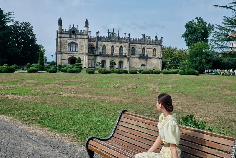 Woman in a pale dress sits on a wooden bench in a quiet park, facing a grand historic castle with trimmed shrubs and expansive green lawn.