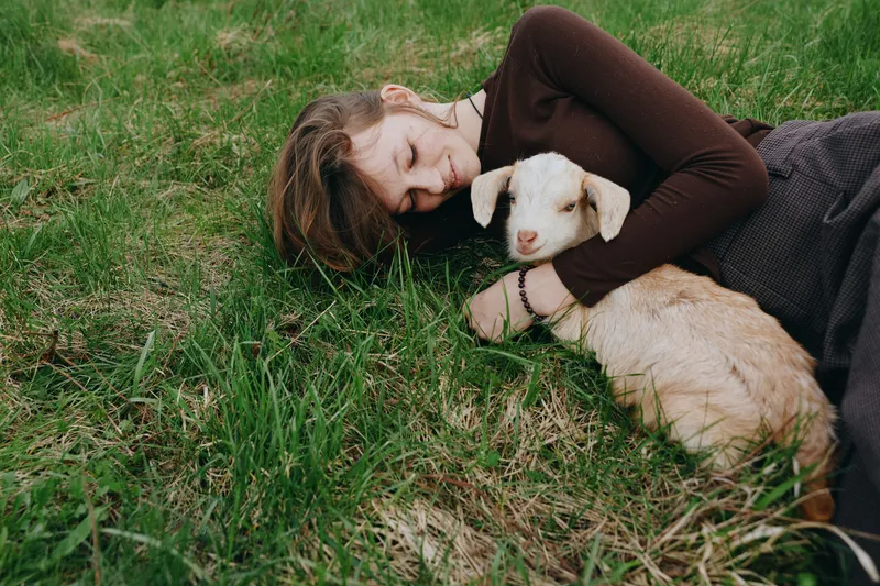 Young woman with long hair lies on green grass embracing a small white goat in a calm outdoor setting, expressing tenderness and connection with nature in a lifestyle moment.
