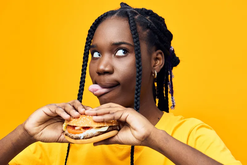 Young Black woman with braided hair playfully sticking her tongue out while holding a delicious burger, vibrant yellow background, expressing fun and enjoyment