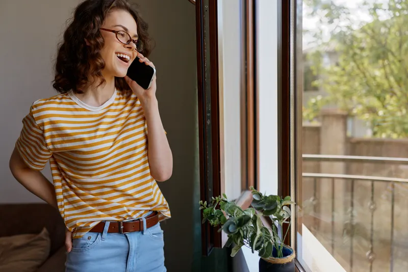 Smiling woman with curly hair wearing a striped shirt stands by a sunlit window, talking on a phone in a cozy home setting. A relaxed lifestyle moment, warm mood, and everyday connectivity.